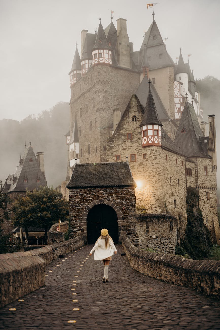 Eltz Castle under cloudy skies. A brown stone castle with pointed half-timbered towers, with a woman walking towards the castle entrance.